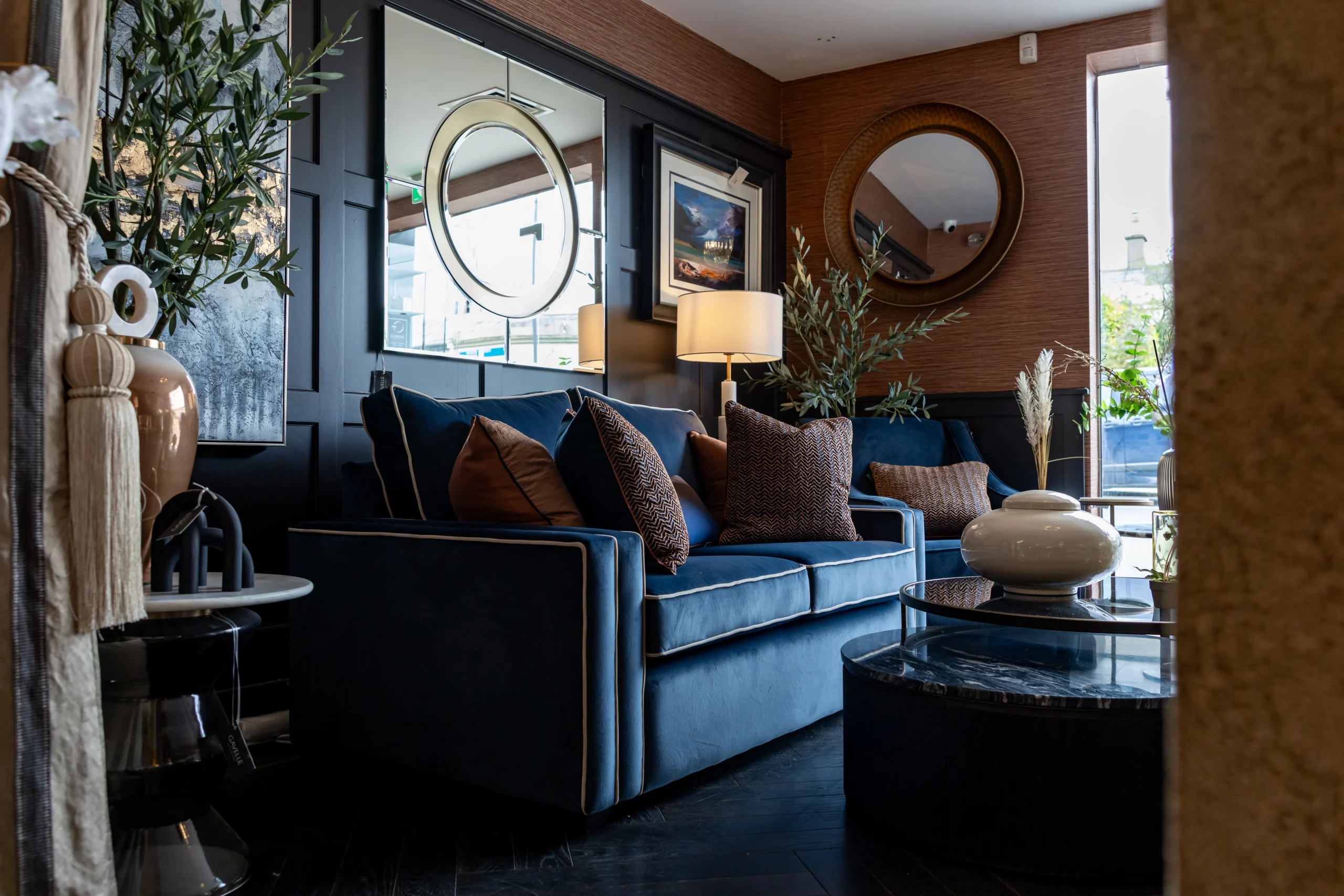 Navy blue velvet sofa with rust brown cushions, round gold mirror and black panelled wall