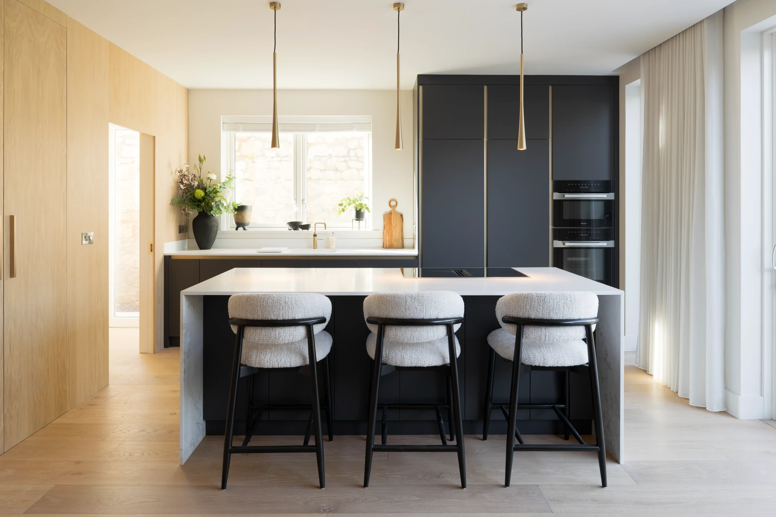 Matte black and oak kitchen with white quartz island, tapered brass pendants and bouclé bar stools