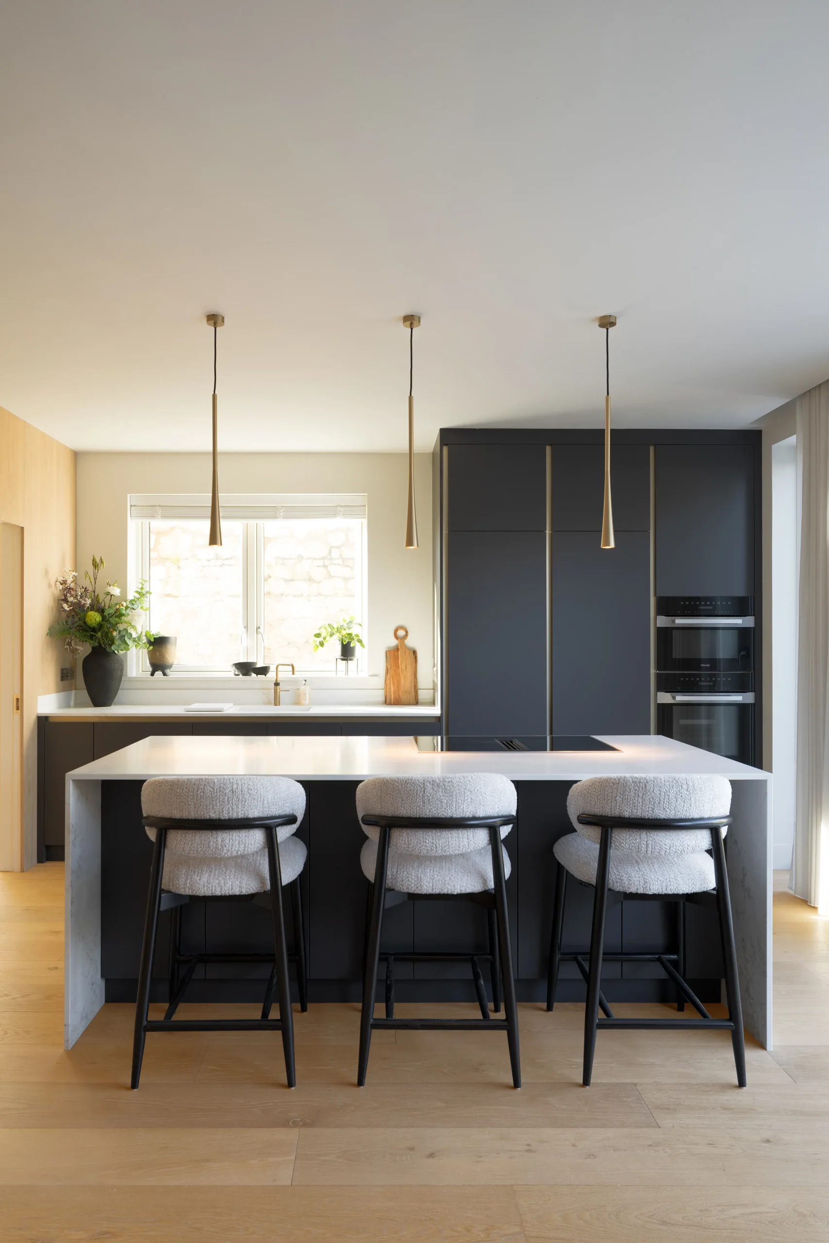 Kitchen island with three cream bouclé bar stools beneath tapered brass pendant lights