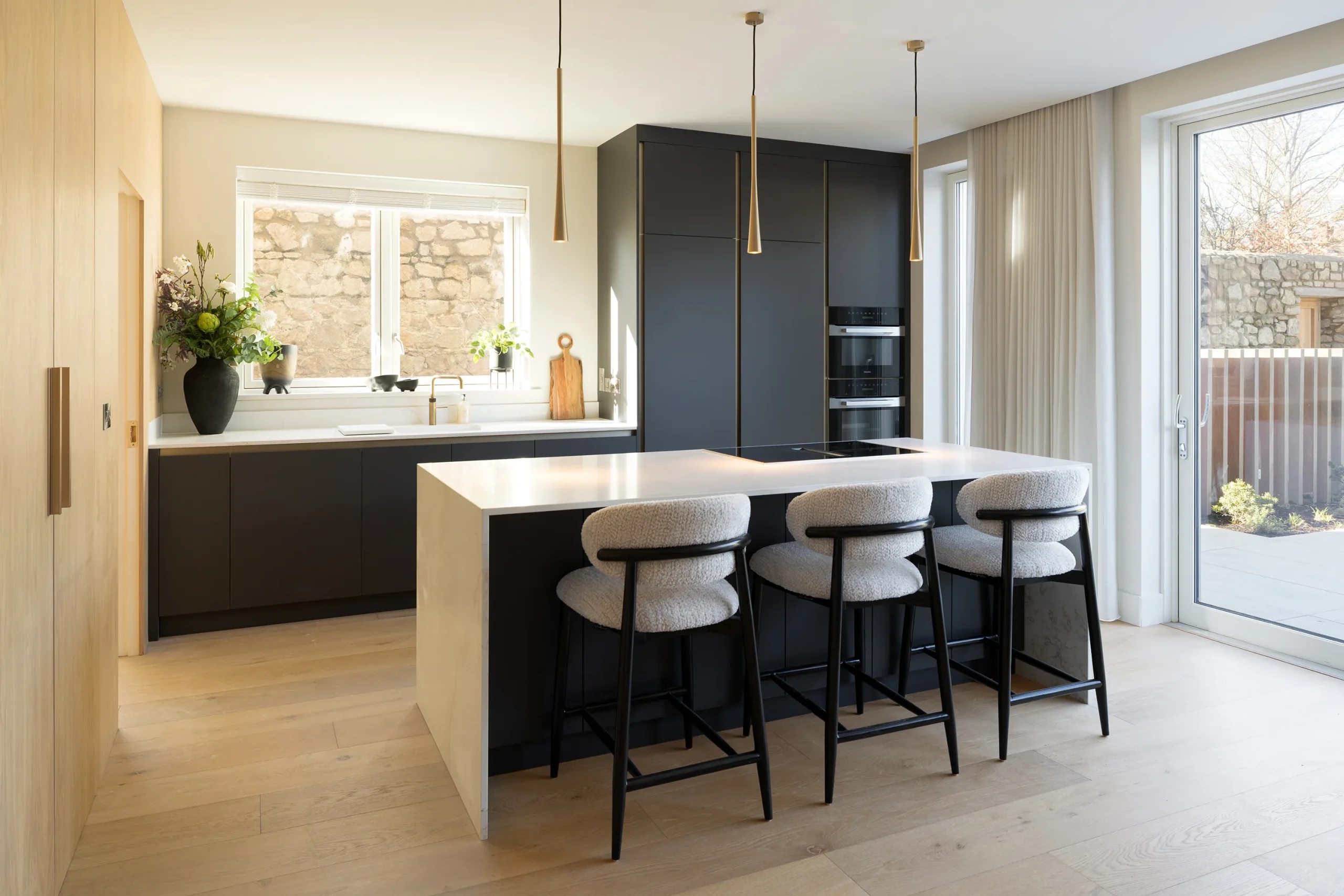 Matte black kitchen with quartz island, cream bouclé bar stools, oak side wall and garden doors to a stone-walled courtyard